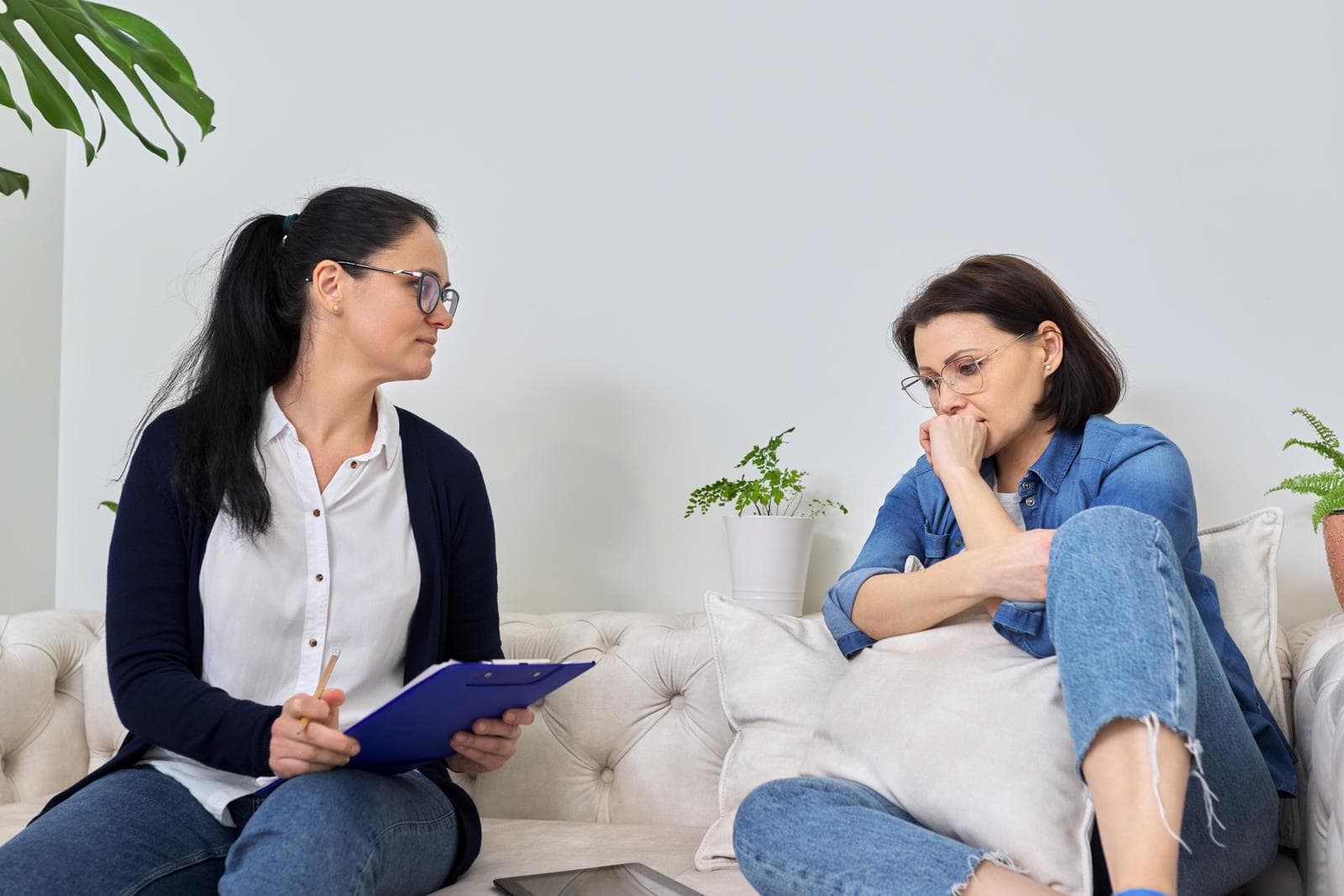 A woman holding a clipboard listens attentively while another woman sits on a couch, looking distressed and holding a pillow.