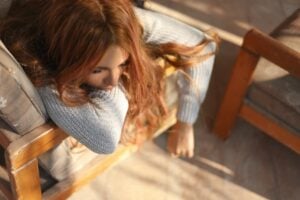 A person with long, wavy brown hair rests their head on the armrest of a gray cushioned chair, with sunlight casting shadows around them.