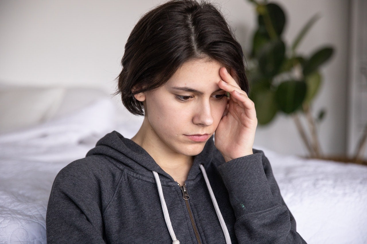 A person with short dark hair wearing a zip-up hoodie looks down while touching their forehead with one hand, appearing deep in thought. A bed and plant are in the background.