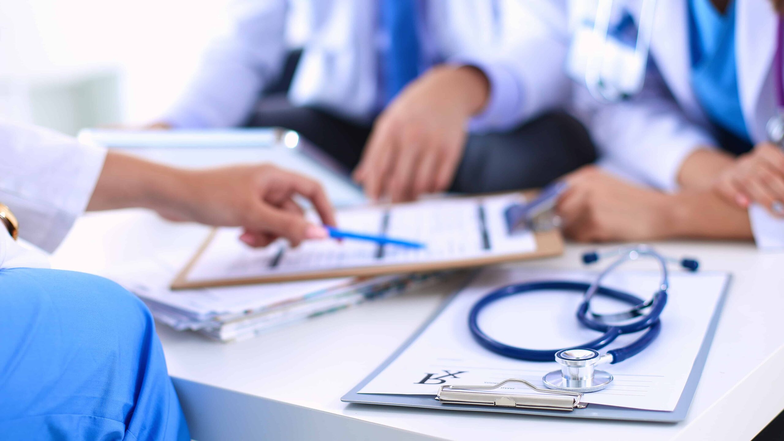 Medical professionals discuss patient charts around a table with a stethoscope and clipboard in the foreground.