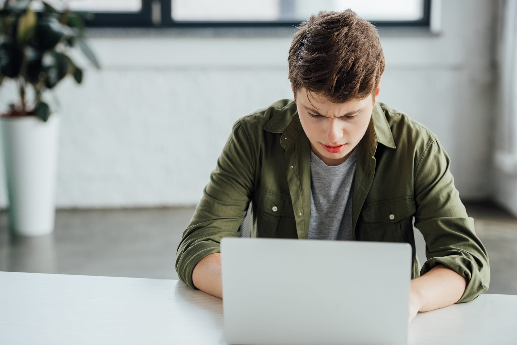 A person with short brown hair wearing a green shirt sits at a table, looking intently at a laptop. A white wall and a potted plant are in the background.