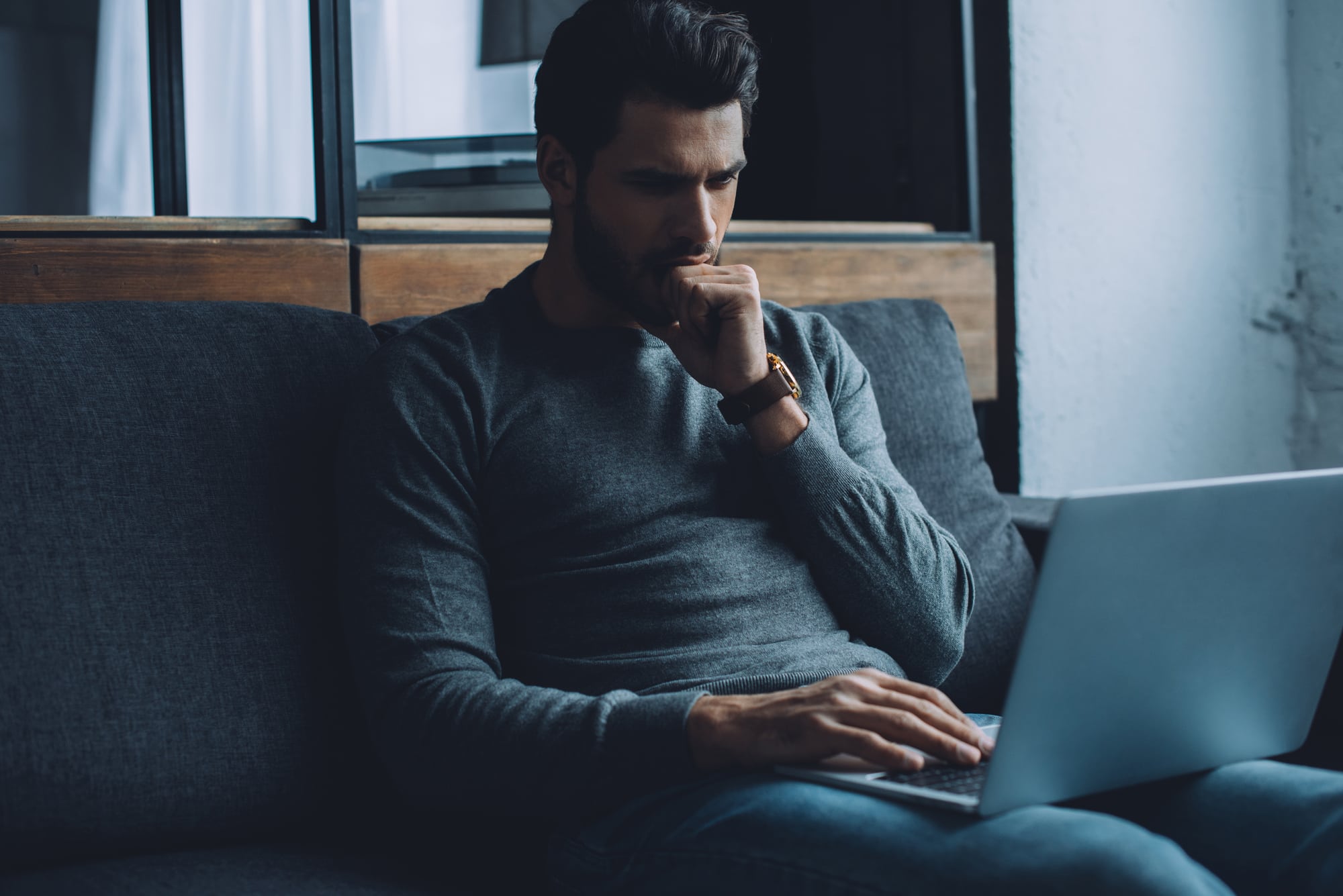 A man sitting on a dark gray couch, wearing a gray sweater, is thoughtfully looking at a laptop on his lap with his right hand near his chin.